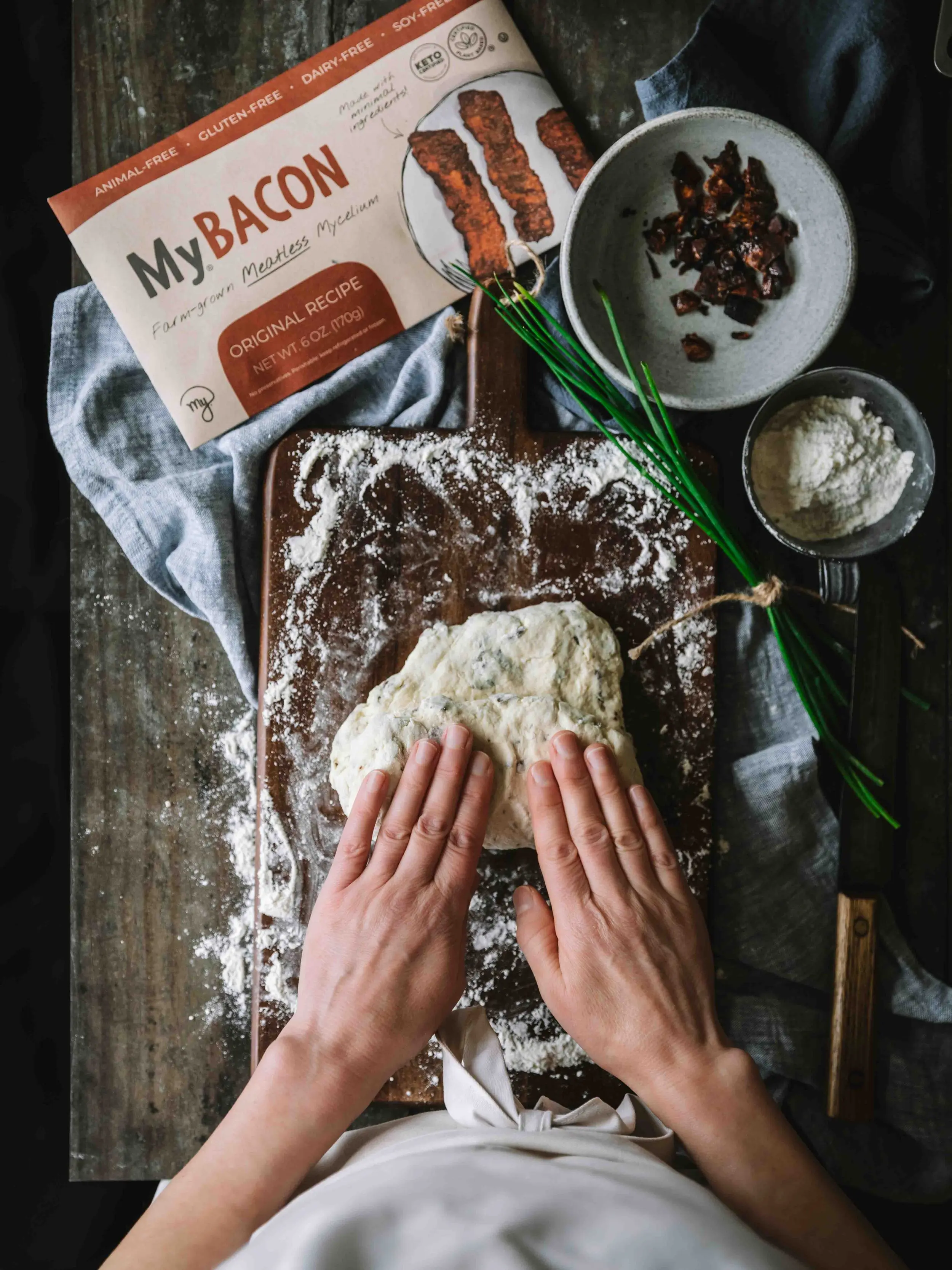 Two hands folding the MyBacon Biscuit dough into hald and onto intself. Other ingredients are scattered around.
