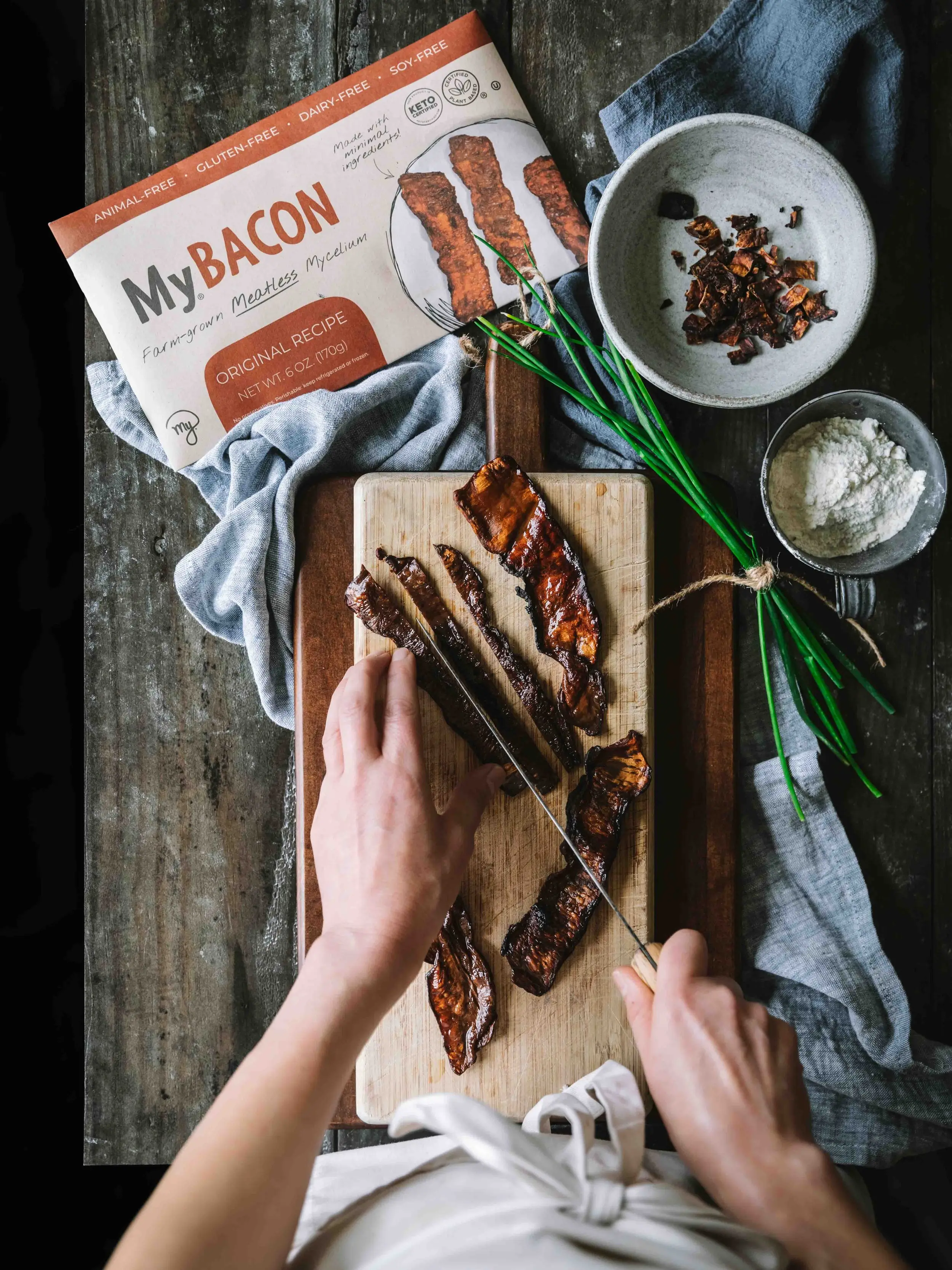 A hand dlicing MyBacon into 1-inch pieces on a nice wooden cutting board. Other ingredients are scattered about, along with a pack of MyBacon