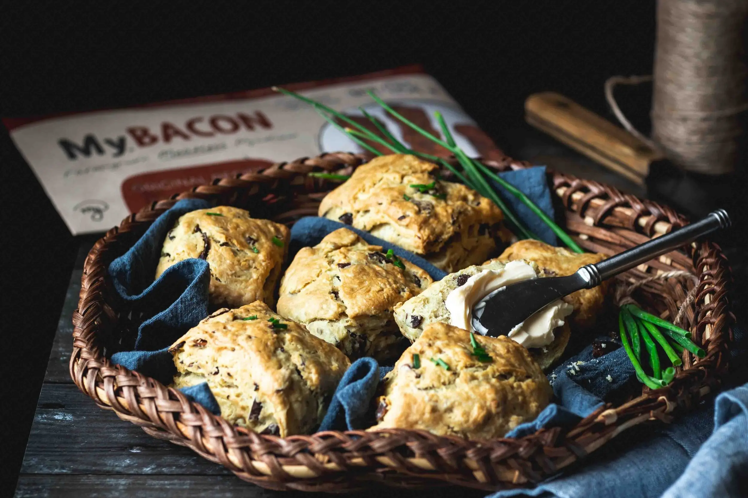 Image of MyBacon Biscuits arranged nicely in a basket. One biscuit is being buttered by a silver knife. You can see a pack of MyBacon out of focus in the top left-hand corner