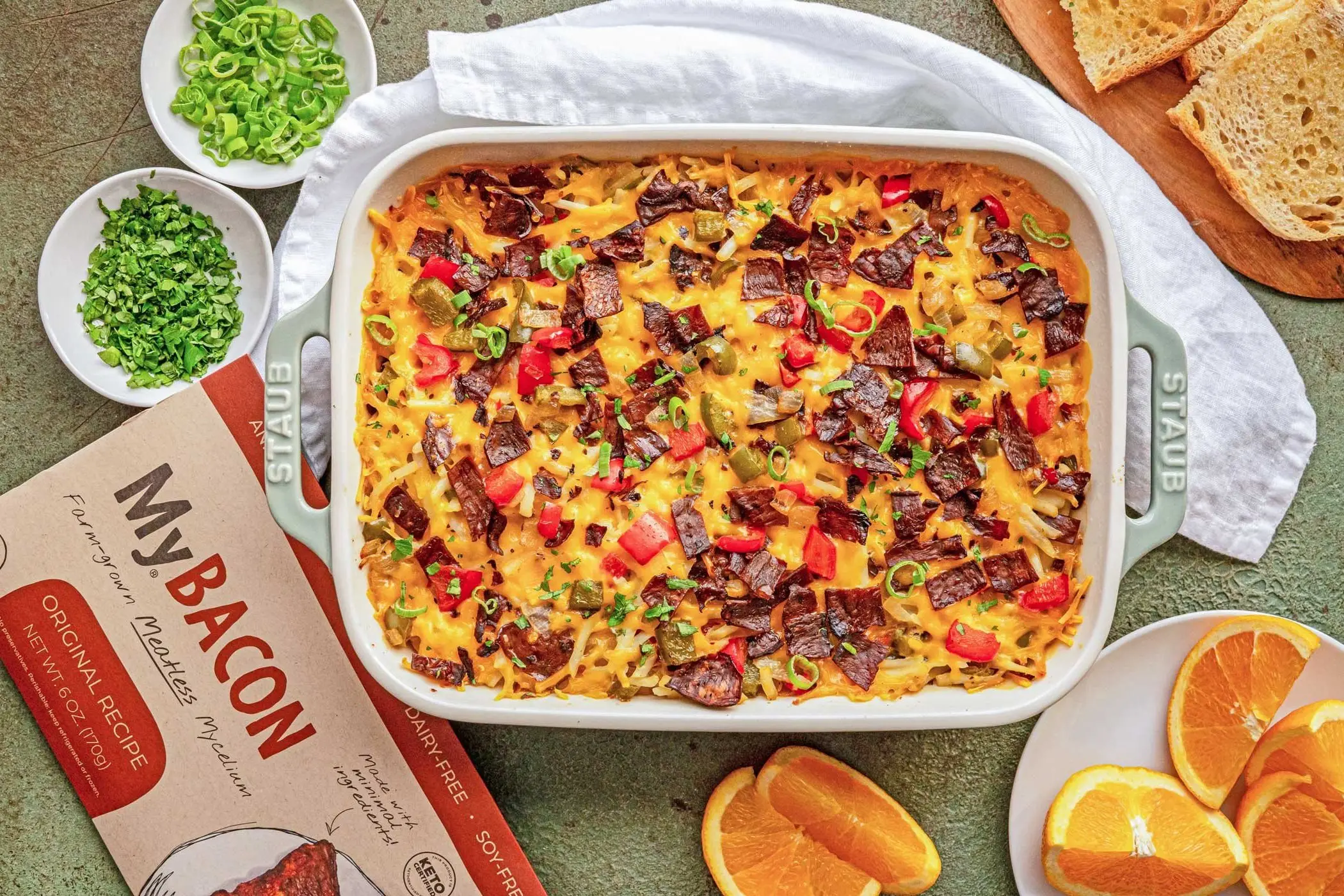 Top-down image of a cooked full pan of MyBacon Breakfast Bake on a green couunter with a white cloth napkin, chopped green onion, chopped parsley, slices of orange and a pack of MyBacon