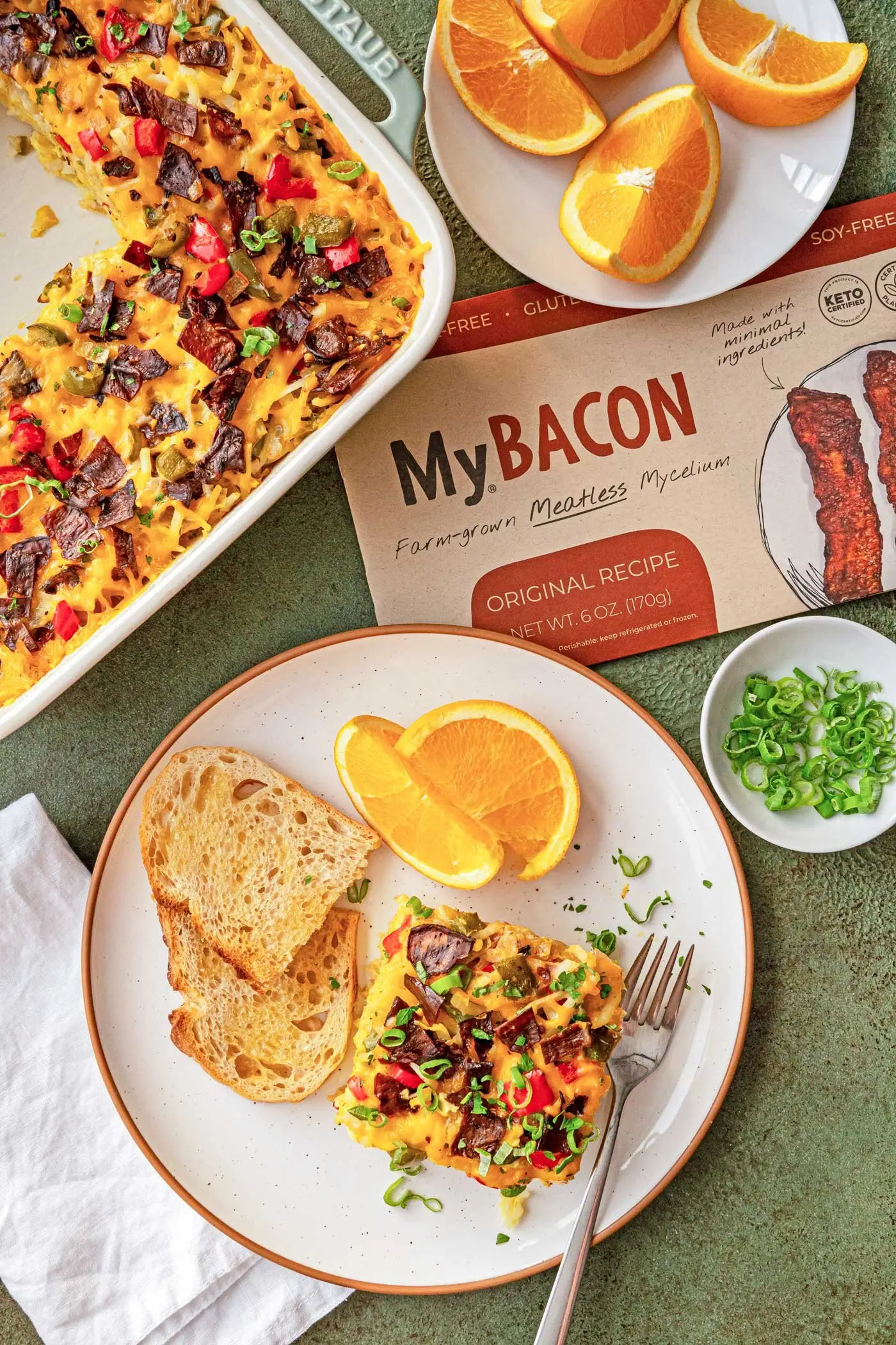 Top down shot of baking dish filled with MyBacon Breafkast Bake, a pack of MyBacon and a plate containing a slice of MyBacon Breakfast Bake with toast, orange slices and a fork.