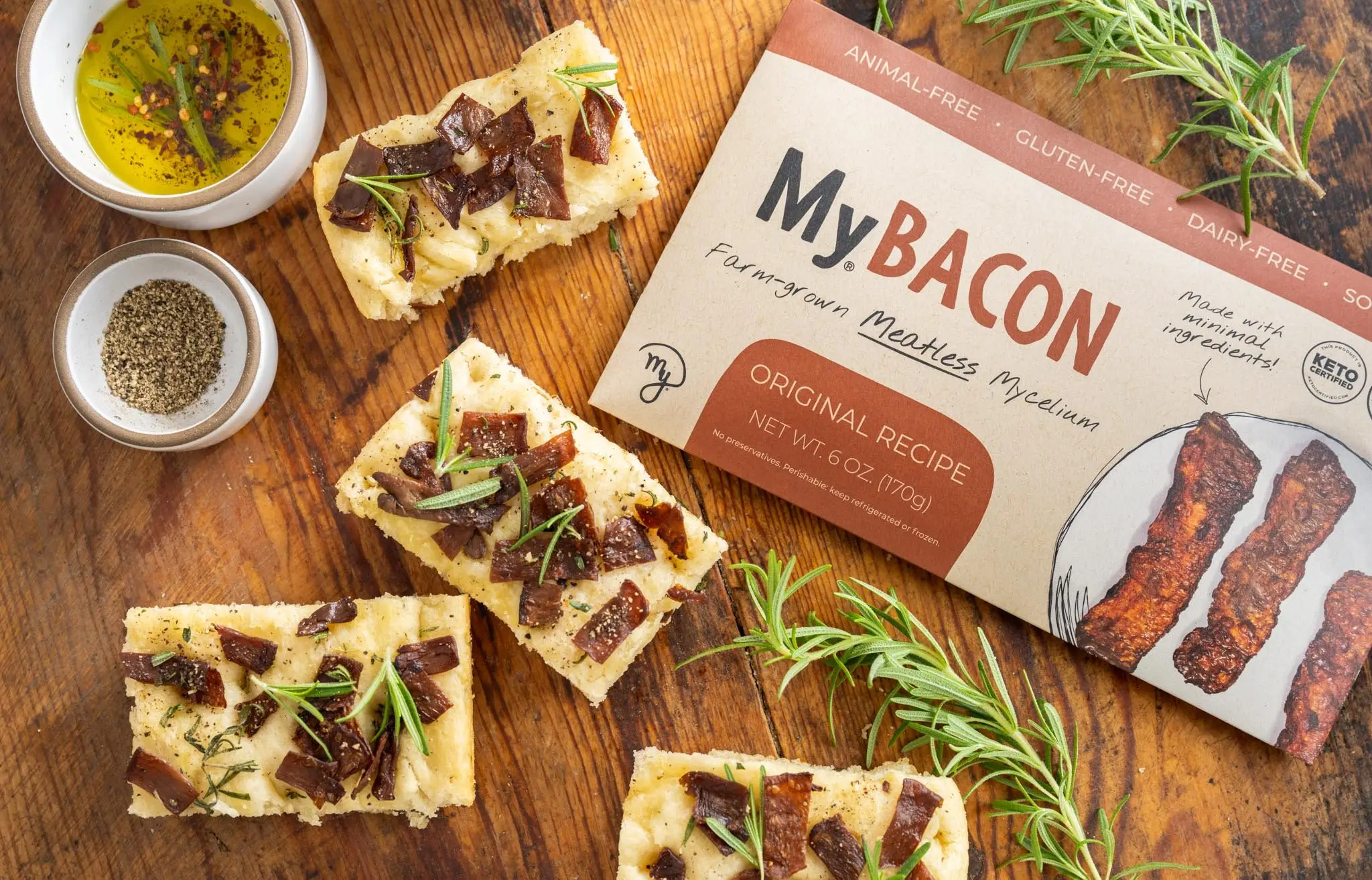 A Top-down photo of 4 slices of Rosemary MyBacon Focaccia on a wooden table with springs of rosemary, a bowl of pepper, bowl of olive oil, and pack of MyBacon.