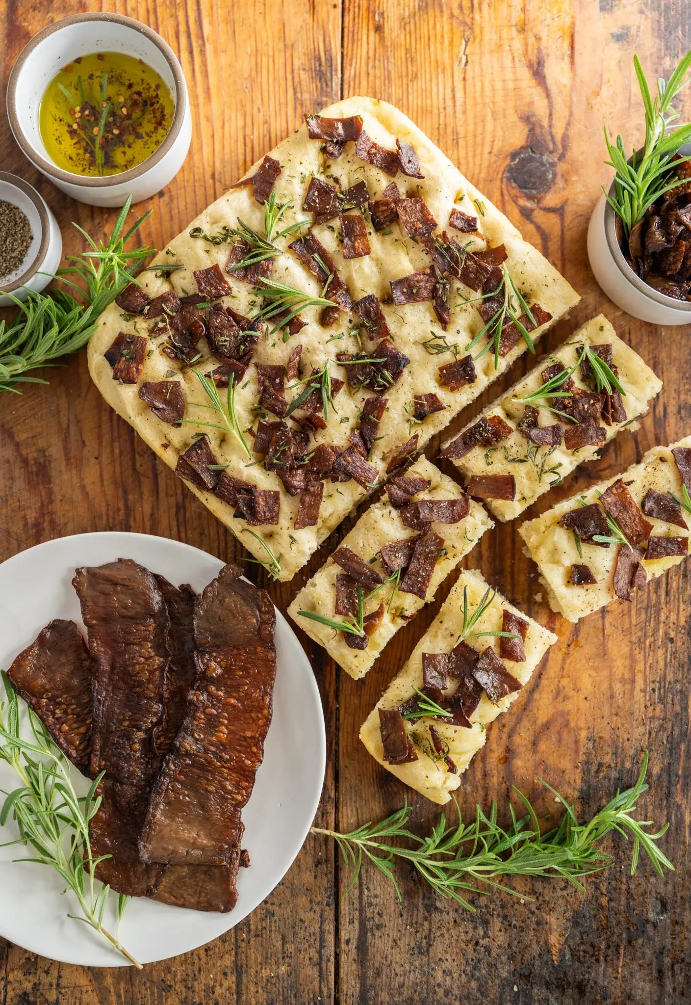 Top-down Photo of a partially sliced loaf of Rosemary MyBacon Focaccia on a wooden table with springs of rosemary and a white plate containing 3 slices of MyBacon