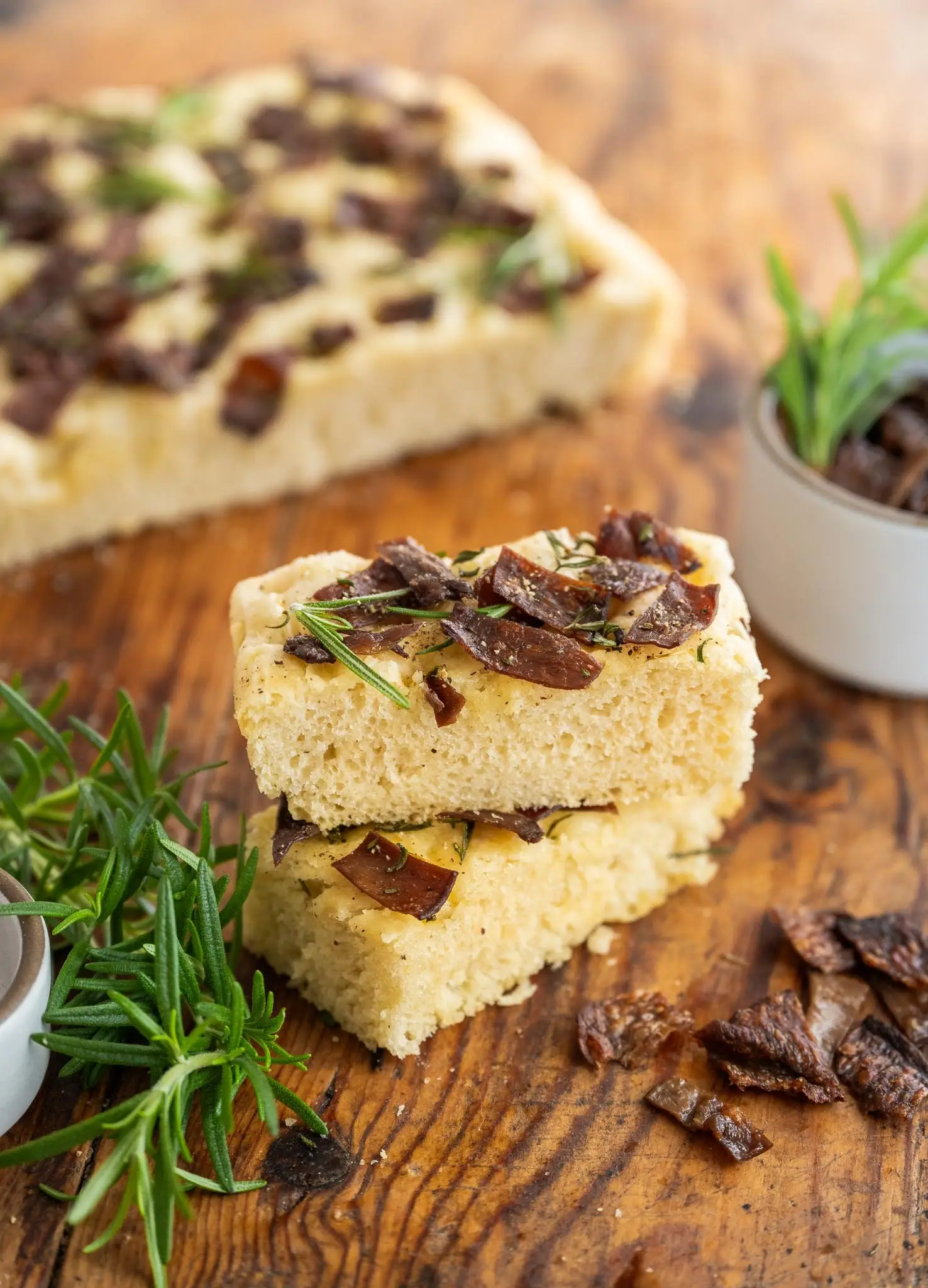 Side shot of two stacked slices of Rosemary MyBacon Focaccia in focus wiht springs of rosemary on wooden table, with blurred out view of full focaccia in the background