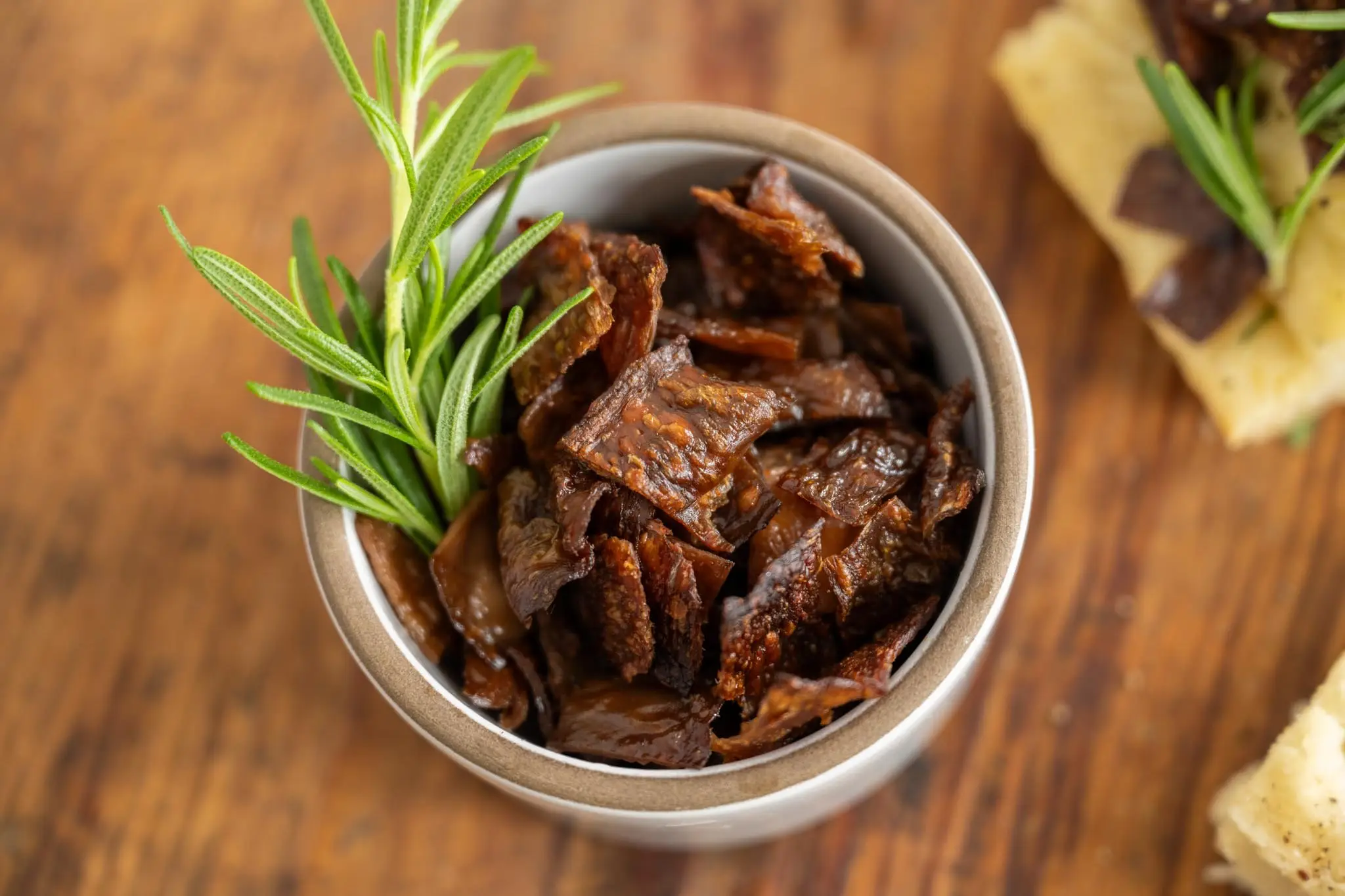 A delicious looking top-down photo of a small ceramic roudn container filled with cut pieces of perfectly cooked MyBacon pieces with a sprig of rosemary sticking out on a wood table